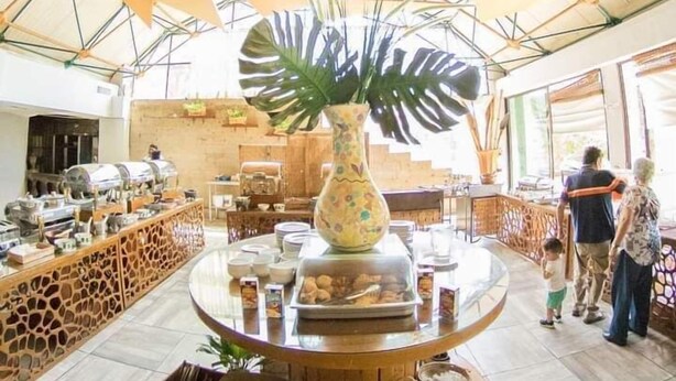 Un vaste espace intérieur doté d’un haut plafond, avec une table ronde centrale entourée de différents récipients contenant de la nourriture et d’une grande plante en pot.