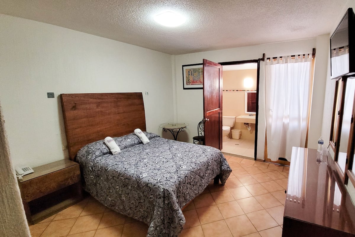 A bedroom with a bed, a bedside table, a small table, a chair, a bathroom with a toilet, and a mirror.