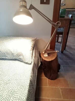 A bed with a patterned bedspread, a desk lamp on a wooden stump, and a kitchen area in the background.