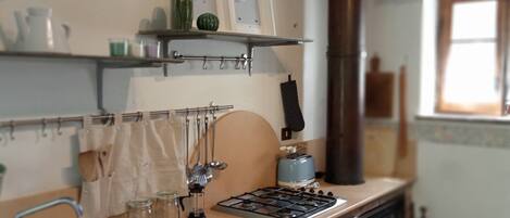 A kitchen with a gas stove, oven, and a sink. There are shelves with kitchen utensils and a window with curtains.