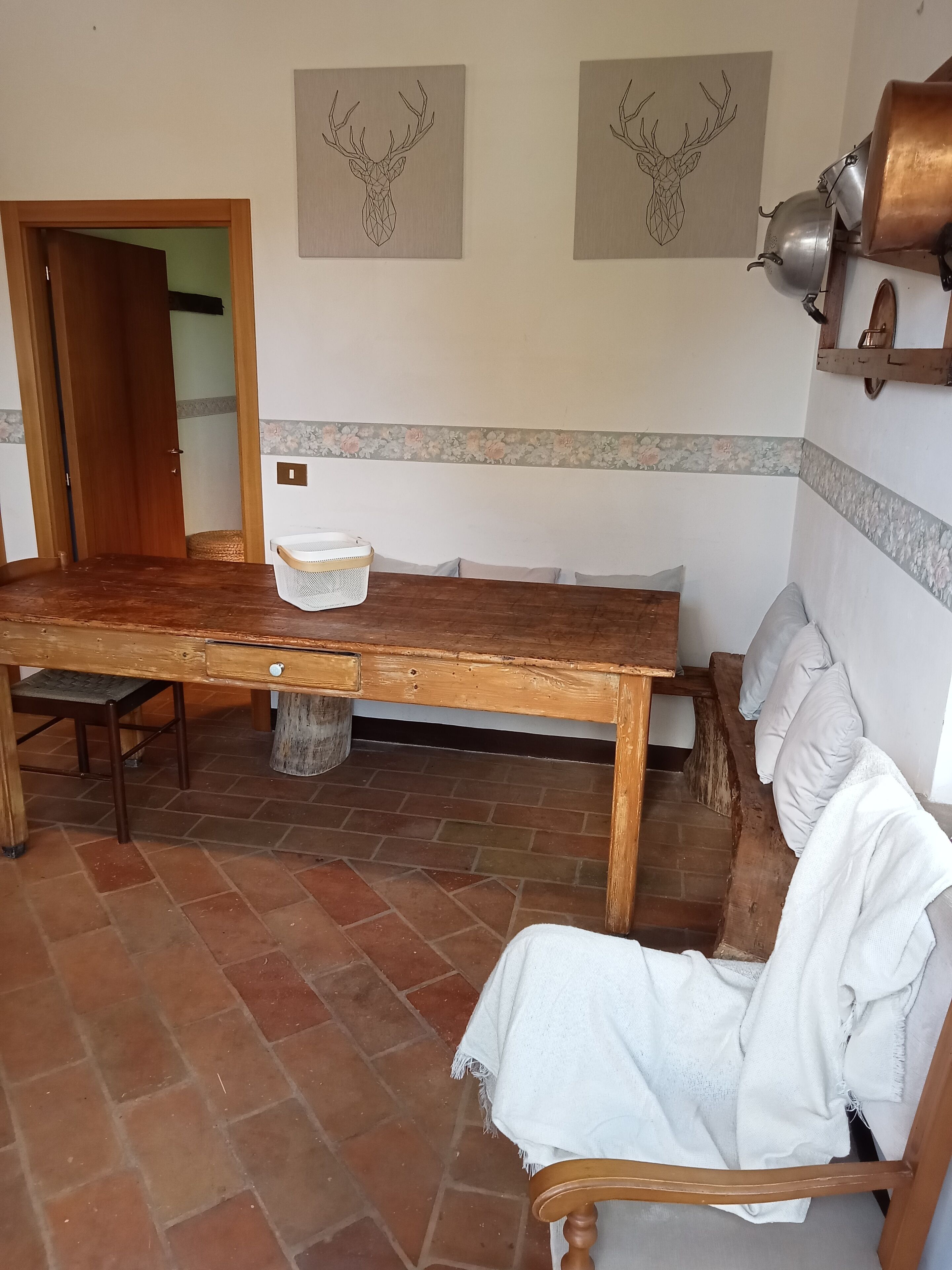 Un comedor rústico con una mesa de madera, sillas y un banco. Hay dos obras de arte colgadas en la pared que representan ciervos. Un mueble de madera con una olla está en la pared, y una cesta sobre la mesa.