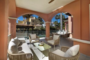 Un espacio de estar techado al aire libre con mobiliario de mimbre y una mesa, con vistas a una piscina y palmeras.
