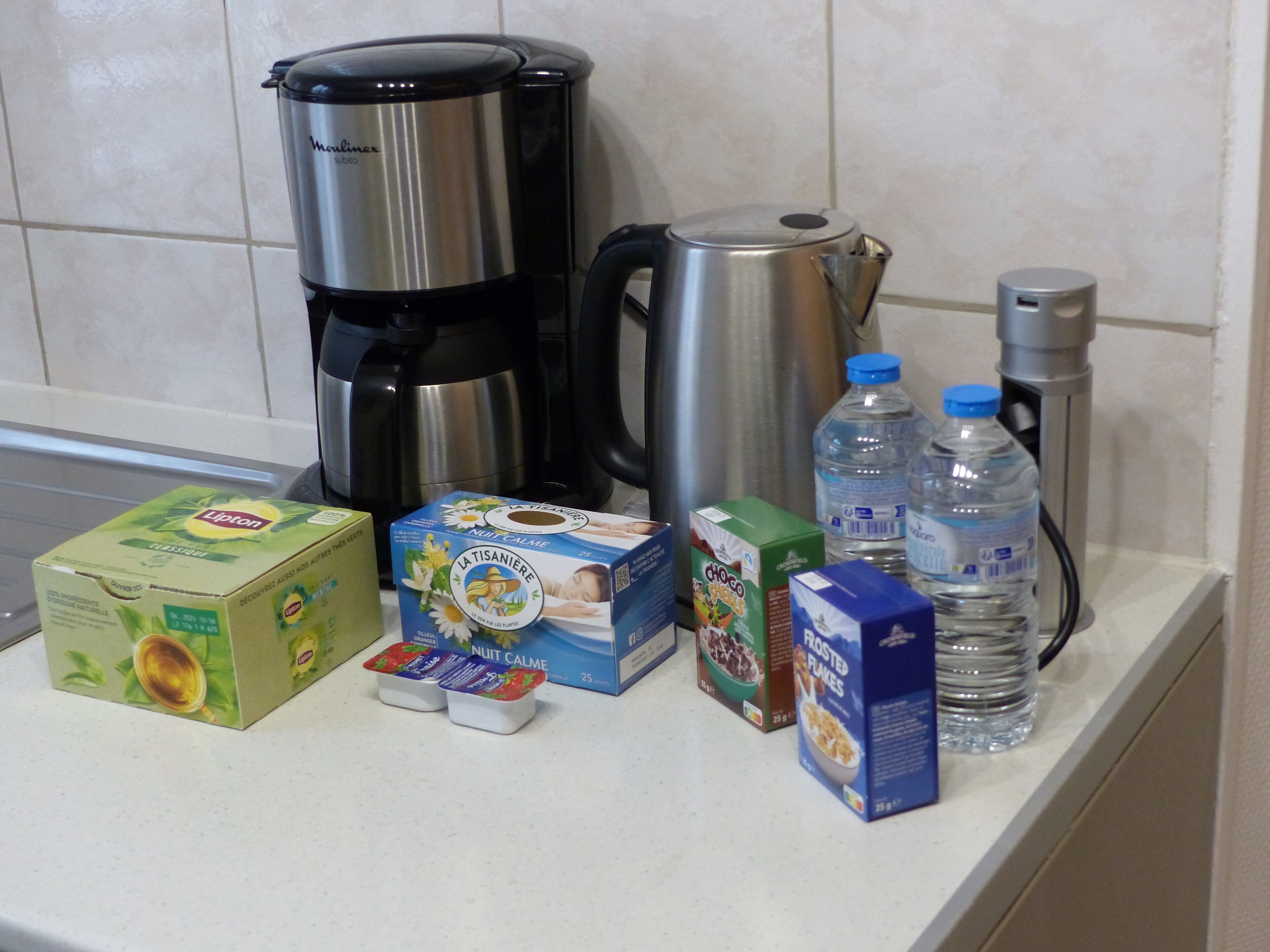 Eine Küchenarbeitsplatte mit Kaffeemaschine, Wasserkocher und verschiedenen Lebensmitteln, darunter Lipton Tee, La Tisaniere Fruchtjoghurt, Choco Flakes Cereal und Wasserflaschen.