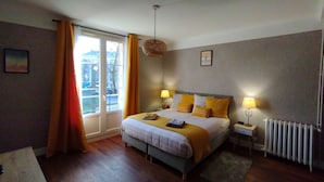 A bedroom with a bed, bedside lamps, a window with curtains, a radiator, and a picture on the wall. - Chez Michèle (Toul)