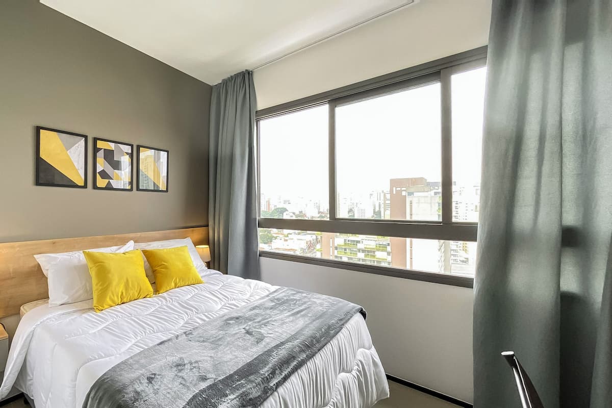 Une chambre d’hôtel moderne avec un grand lit, une table de chevet, une lampe et une fenêtre donnant sur la ville.