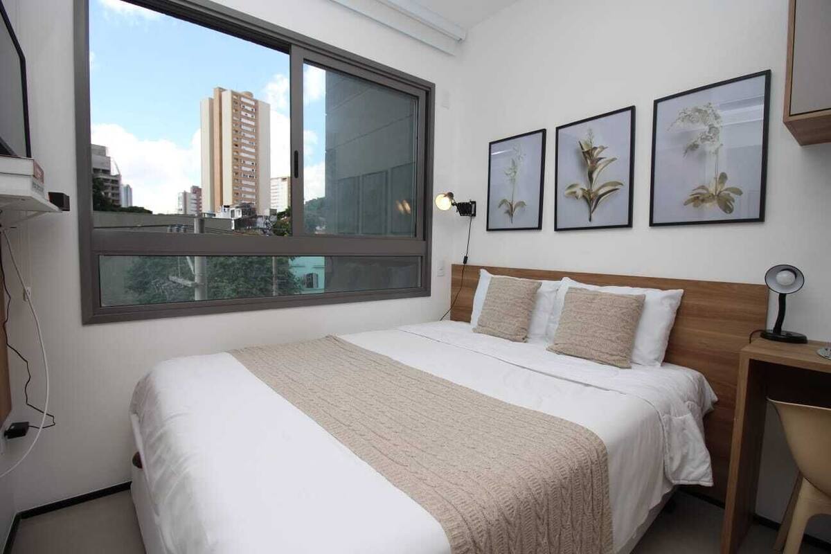 Une chambre avec une grande fenêtre, un lit recouvert d’une literie blanche, une tête de lit en bois et trois tableaux encadrés représentant des motifs floraux accrochés au mur.