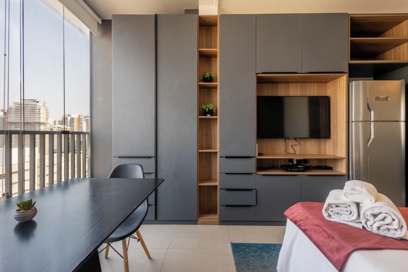 A modern hotel room with a dining table, a TV, a refrigerator, and a bed with towels.