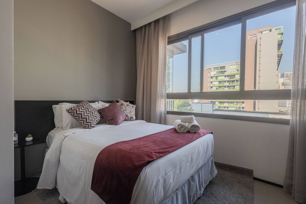 Une chambre d’hôtel avec un lit, une table de chevet, une fenêtre donnant sur des immeubles et un plaid rouge.