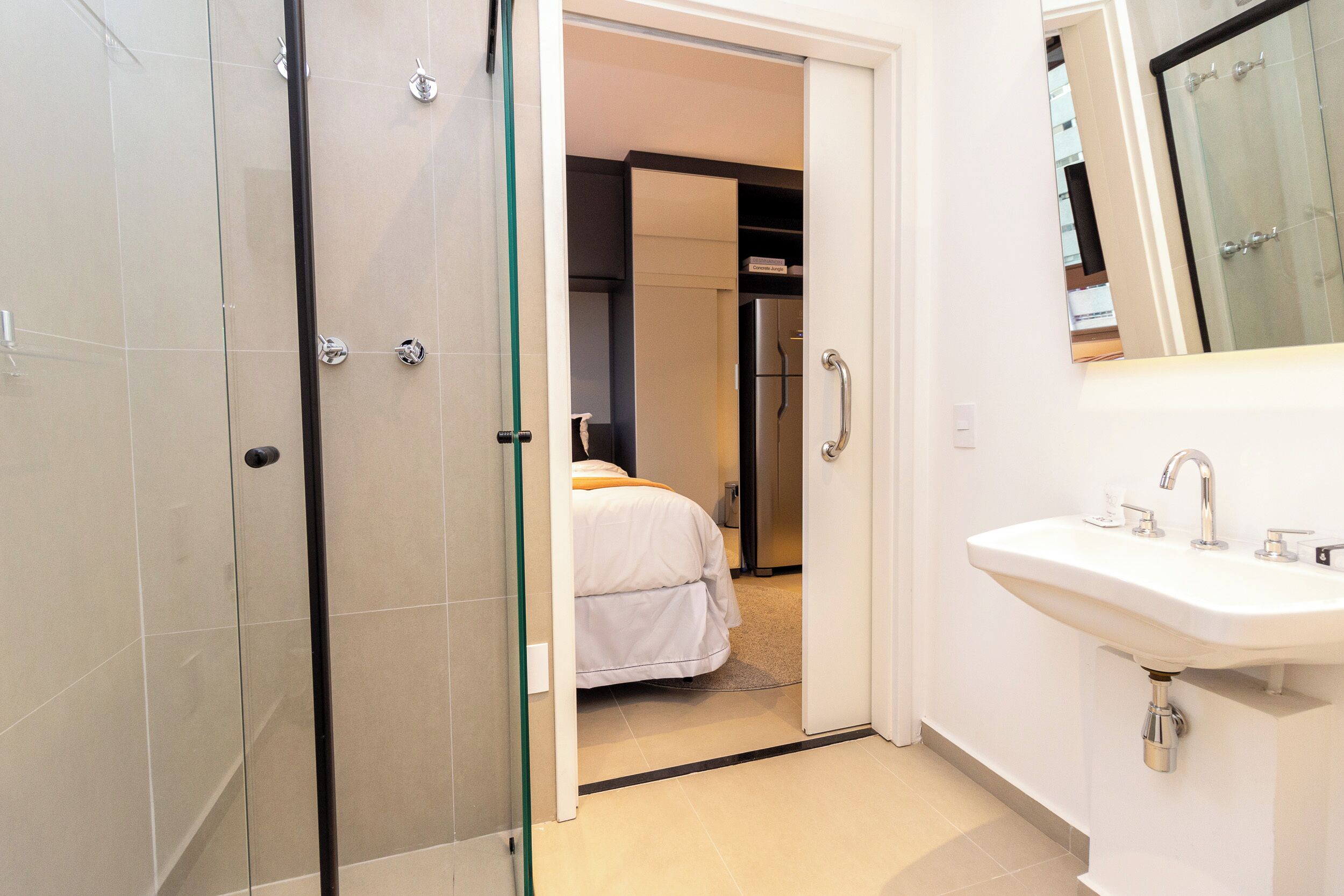 A modern bathroom with a glass shower enclosure, a white sink, and a visible bedroom with a bed and wardrobe.