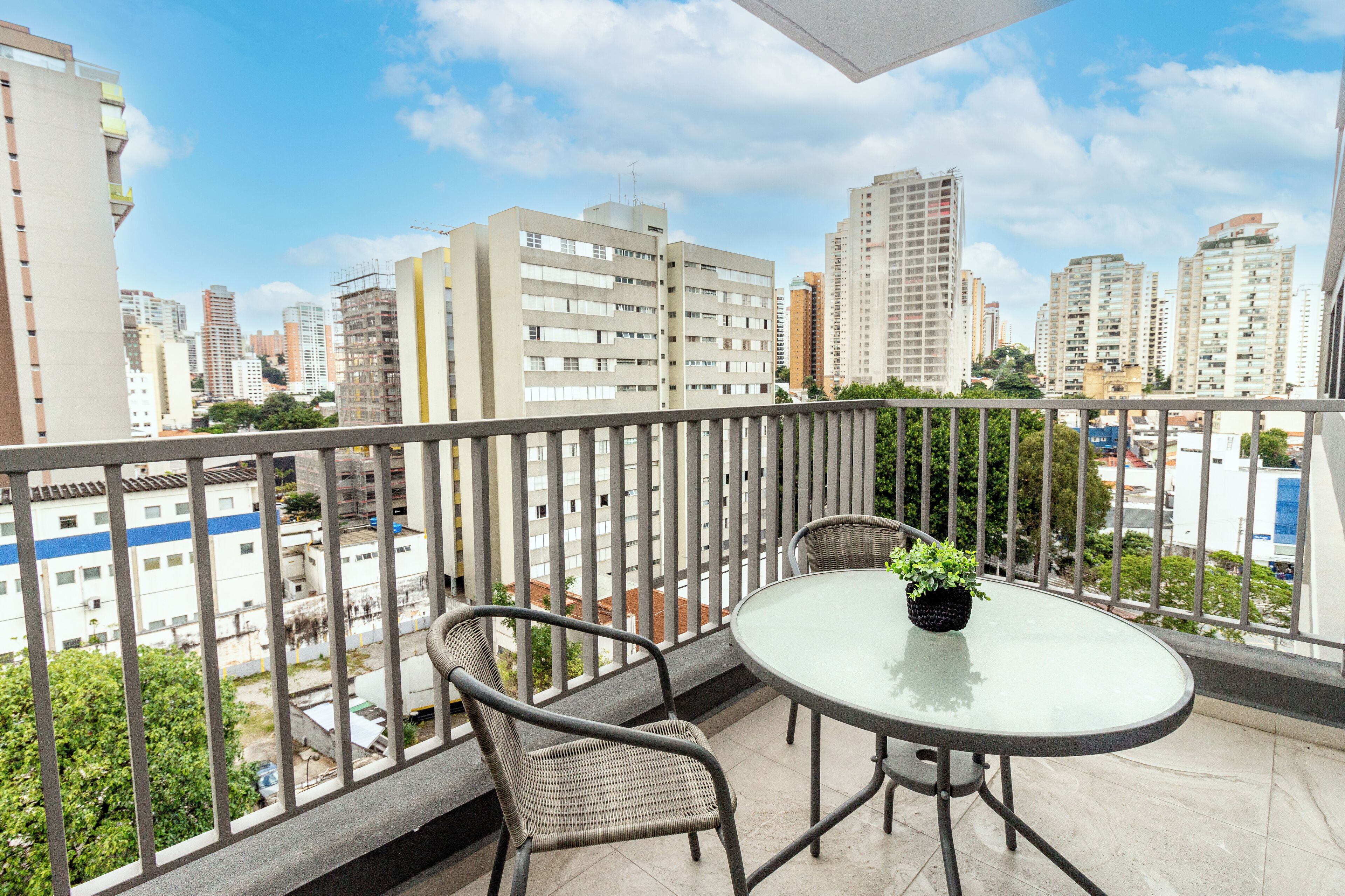 A balcony with a round glass table and two wicker chairs, overlooking a cityscape with high-rise buildings.
