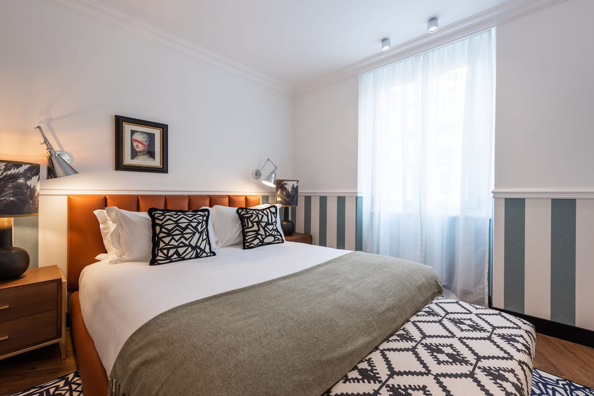 Un dormitorio ordenado con una cama grande, lámparas de noche, un cuadro en la pared y una ventana con cortinas.