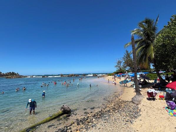 A beach with clear water, people swimming, and umbrellas on the sand.