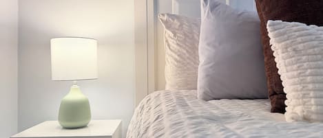 A neatly made bed with white and brown pillows, a white headboard, and a white nightstand with a lamp.