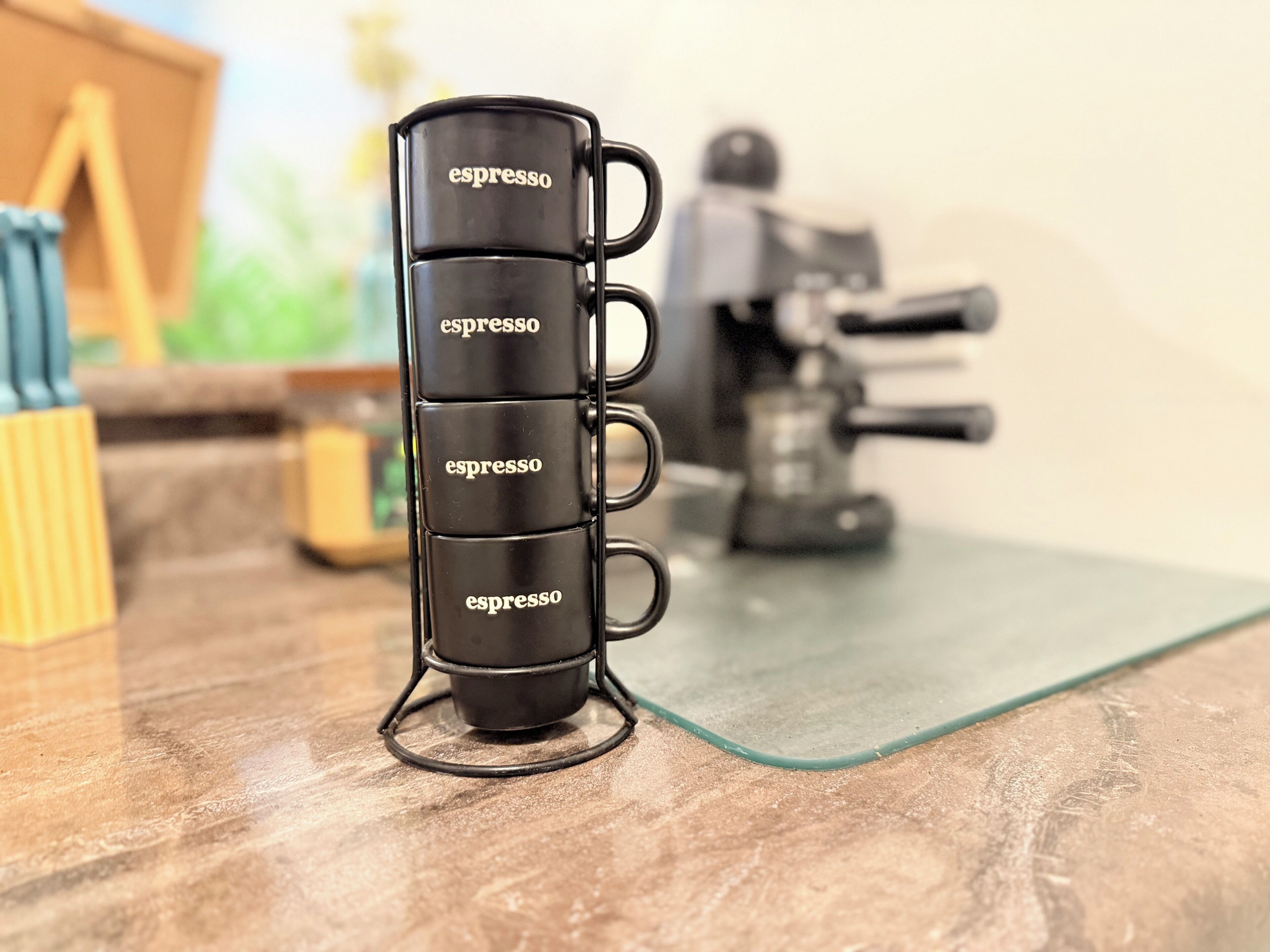 Neljä mustaa espressokuppia on pinottu päällekkäin, ja jokaisessa kupissa on sana »espresso». Kupit on asetettu läpinäkyvälle lasiselle aluslautaselle.