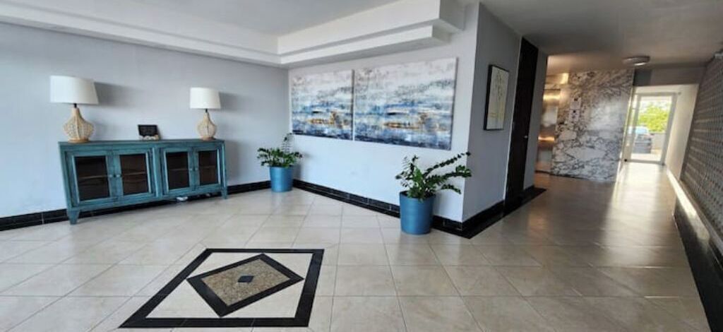 Un hall d’entrée spacieux avec un sol carrelé, une console bleue agrémentée de lampes, des plantes en pot et un grand tableau abstrait accroché au mur.