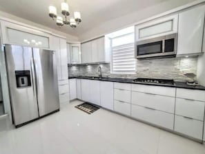 Cozinha moderna com armários brancos, geladeira de aço inoxidável, bancada preta e janela com persianas.
