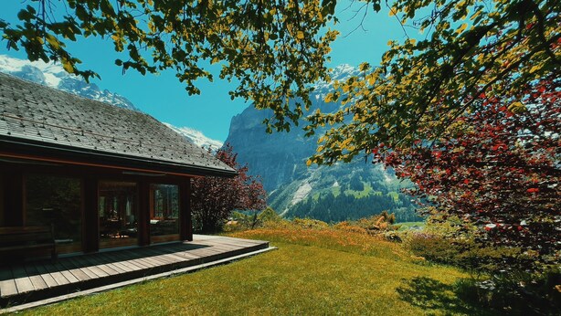 A modern building with a wooden deck and a bench, surrounded by lush greenery and mountains in the background. - Alpine Escape - Iconic Views & Timeless Design (Grindelwald)