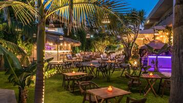 An outdoor patio area with wooden tables and chairs, surrounded by palm trees and greenery.