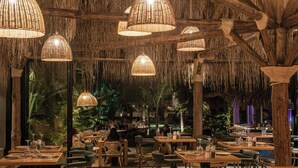 An indoor restaurant with wooden tables and chairs, hanging wicker lamps, and a tropical ambiance with plants and a thatched ceiling.