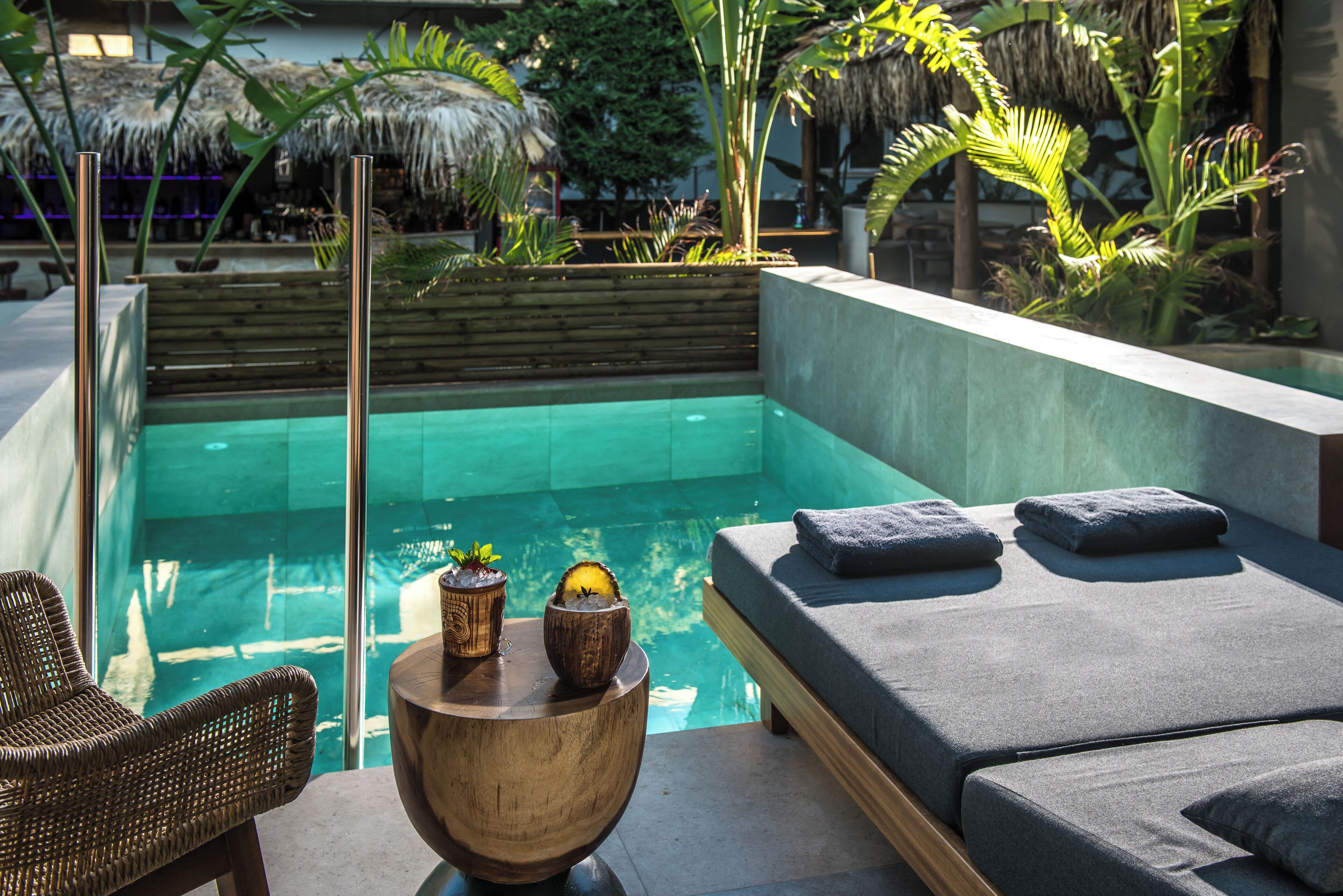 A modern outdoor pool area with a lounge chair, a wooden side table with decorative items, and a cushioned seating area with towels.
