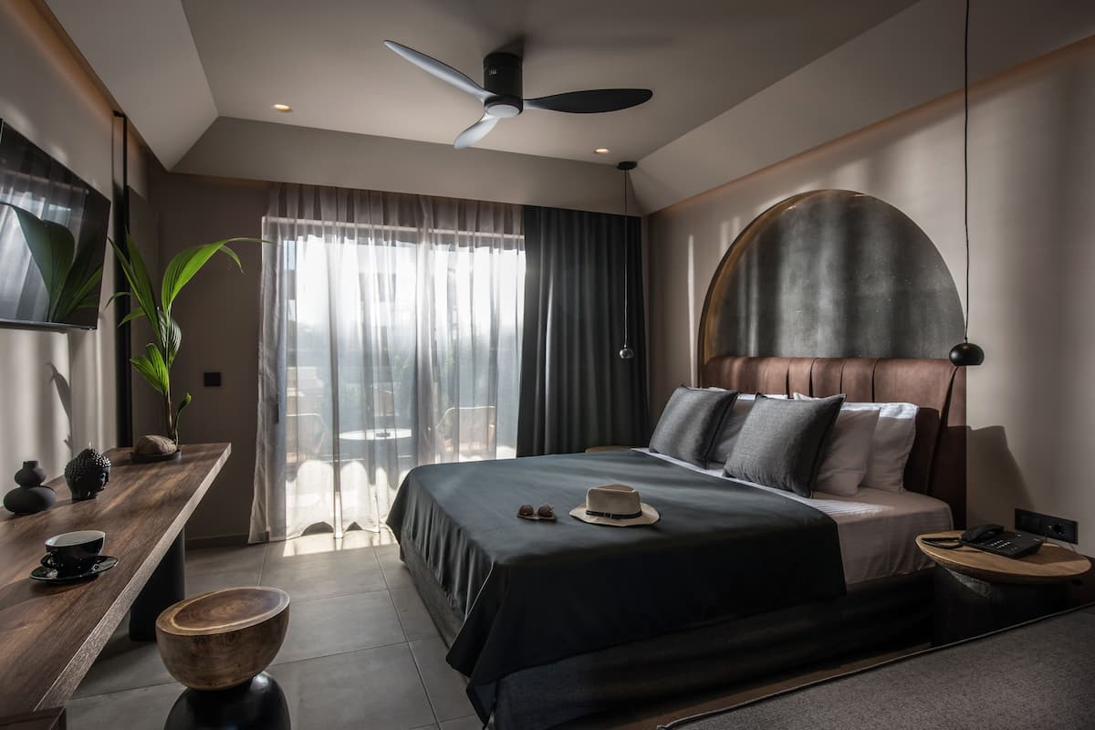 Ett modernt hotellrum med en stor säng, ett skrivbord med en skål, en takfläkt och ett stort fönster med gardiner.
