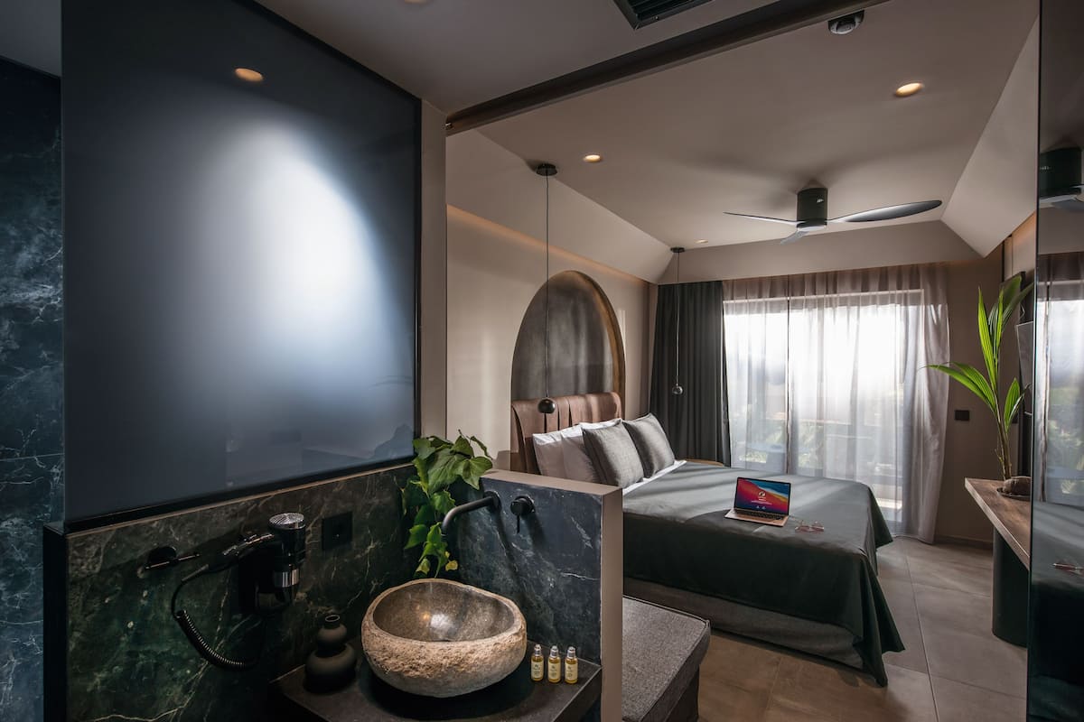 Ett modernt hotellrum med en stor säng, ett inbyggt skrivbord, ett stort fönster med gardiner, ett handfat i marmor och en växt.