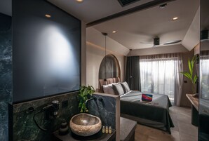 A modern hotel room with a large bed, a built-in desk, a large window with curtains, a marble sink, and a plant.