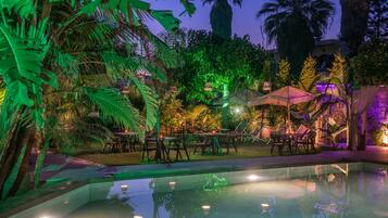 An outdoor pool area with lounge chairs and umbrellas, surrounded by lush tropical plants and palm trees.
