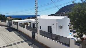 A modern building with a white facade, a metal fence, and a view of the sea and a mountain in the background. - Modern luxury new build villa paradise, sea view, aircon, pool (Calpe)