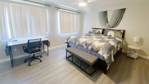 Een slaapkamer met een bed, een bureau, een stoel, een bankje, een staande lamp en een plafondventilator.