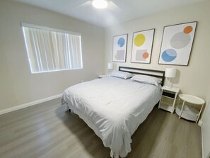 A bedroom with a bed, a nightstand, a bedside table, a window with blinds, and three framed abstract art pieces on the wall.