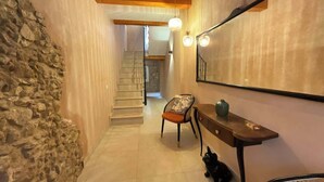 Un couloir étroit, avec un mur en pierre d’un côté et un plafond avec des poutres en bois. Une chaise et une petite table sont placées contre le mur, et un miroir est accroché au-dessus. On aperçoit un escalier au fond.