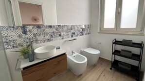 A modern bathroom with a white sink, bidet, and toilet, a decorative backsplash, and a black storage cart. - RUA 39 fine apartments 4 (Syracuse)
