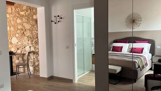 A modern bedroom with a large bed, a bedside table, and a chair. There is a stone wall and a glass door leading to another room. - RUA 39 fine apartments 4 (Syracuse)