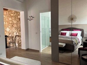 Une chambre moderne avec un grand lit, une table de chevet et une chaise. On y trouve un mur en pierre et une porte en verre qui donne sur une autre pièce.