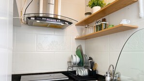Une cuisine équipée d’un évier en acier inoxydable, d’un égouttoir et d’une cuisinière. Au-dessus du plan de travail, on trouve des étagères en bois où sont rangés des ustensiles de cuisine et une plante en pot.