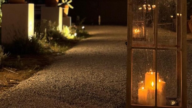 Un sendero de grava conduce a una casa con un farol y velas, rodeada de plantas en macetas y un cielo al atardecer.