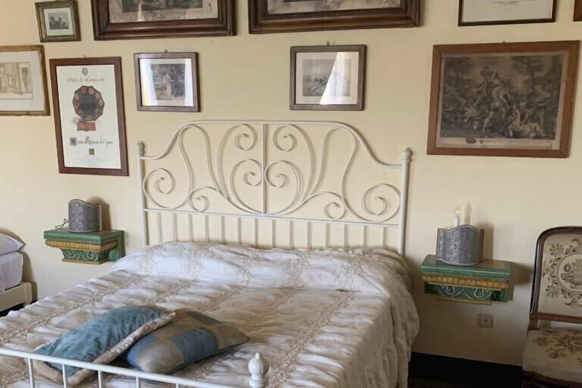 A bedroom with a white metal bed frame, a patterned bedspread, and a blue pillow. There are framed pictures on the wall, a green nightstand with a lamp, and a patterned rug on the floor.