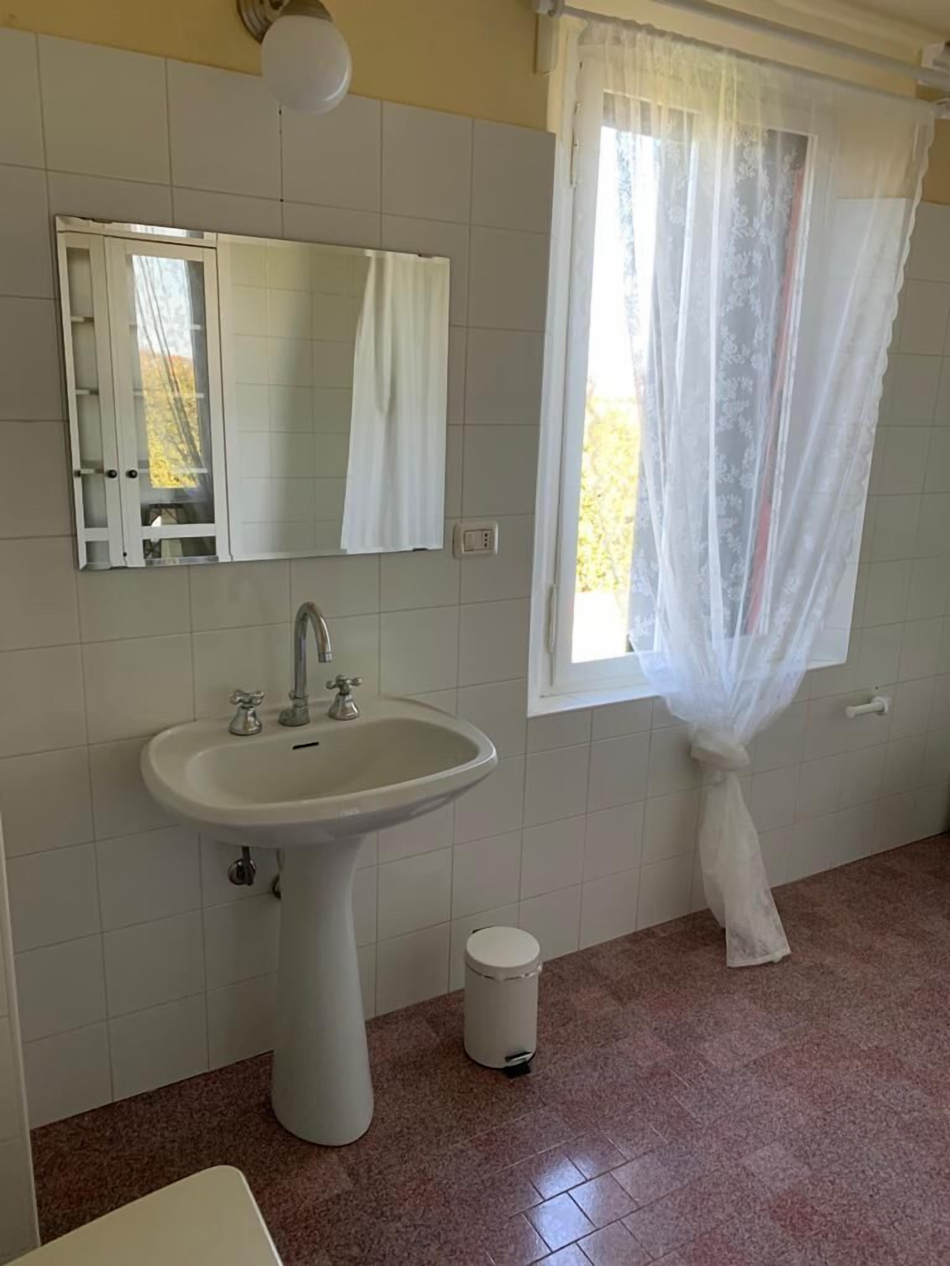 Un baño con un lavamanos pedestal, un espejo, una ventana con cortina translúcida y un cubo de basura.