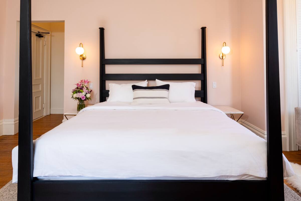 A neatly made bed with white linens and a black headboard in a room with light pink walls and two wall-mounted light fixtures.