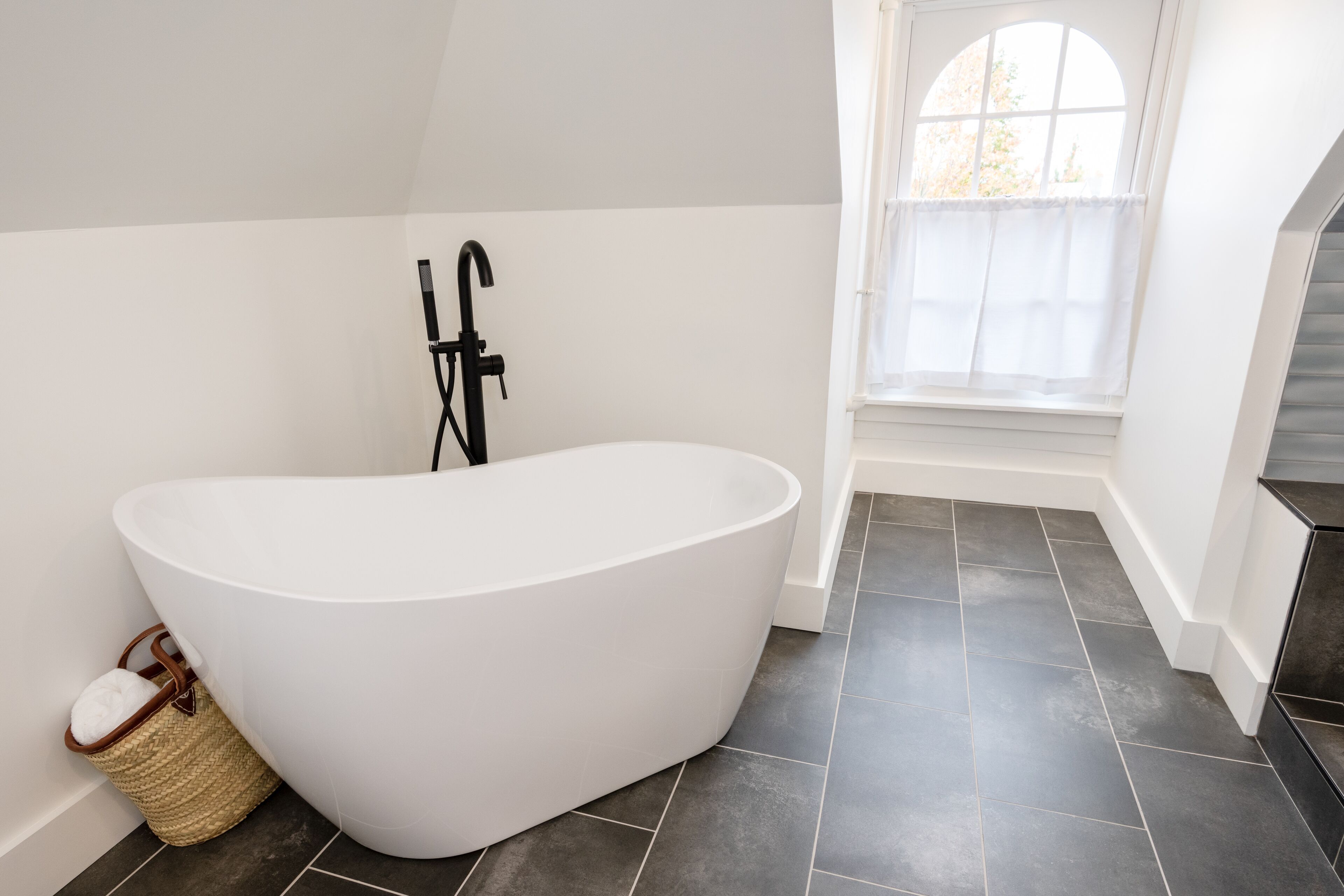 A modern bathroom with a freestanding bathtub, a window with a view, and dark tiled flooring.
