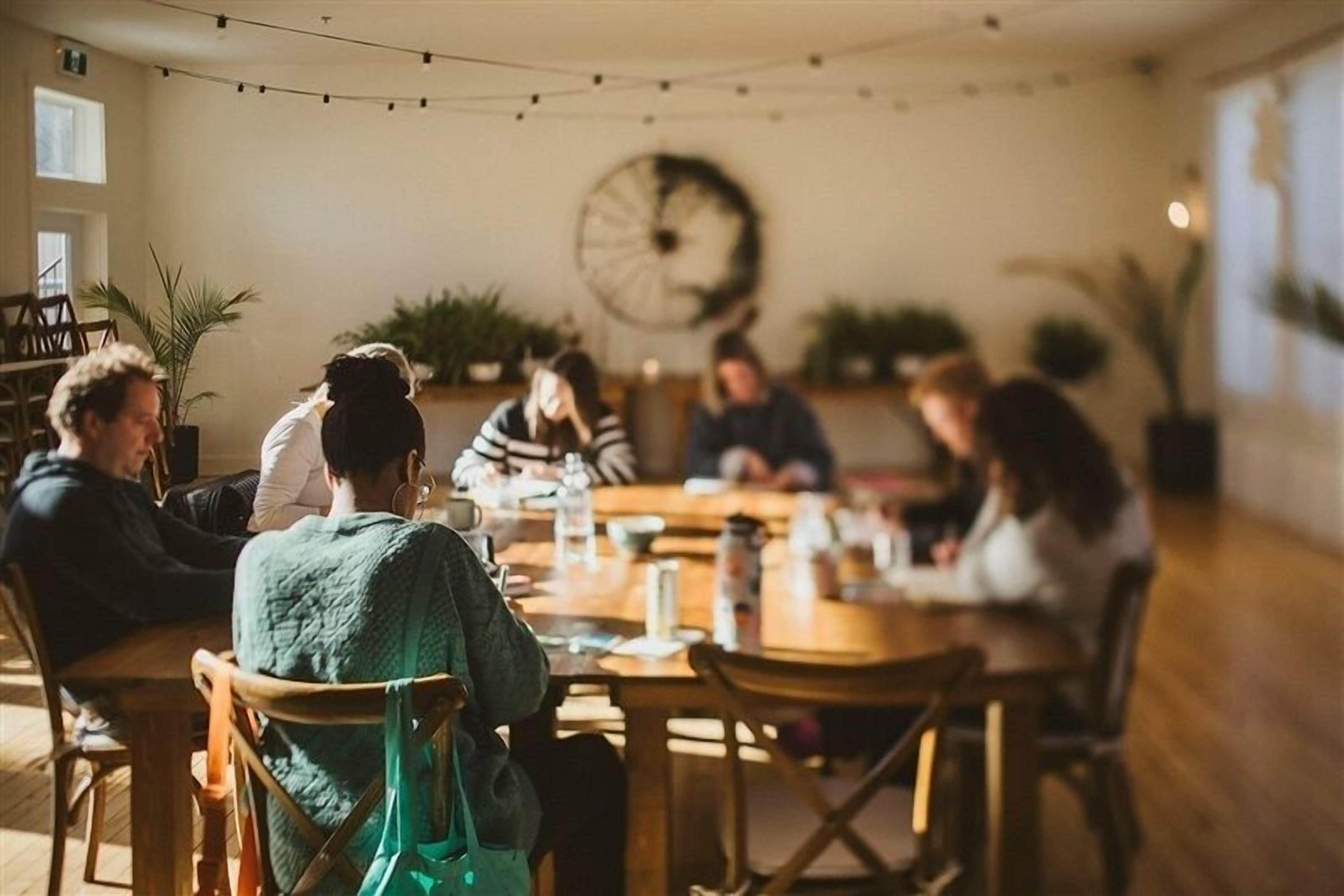 A group of people seated around a wooden table in a room with plants and a clock on the wall.