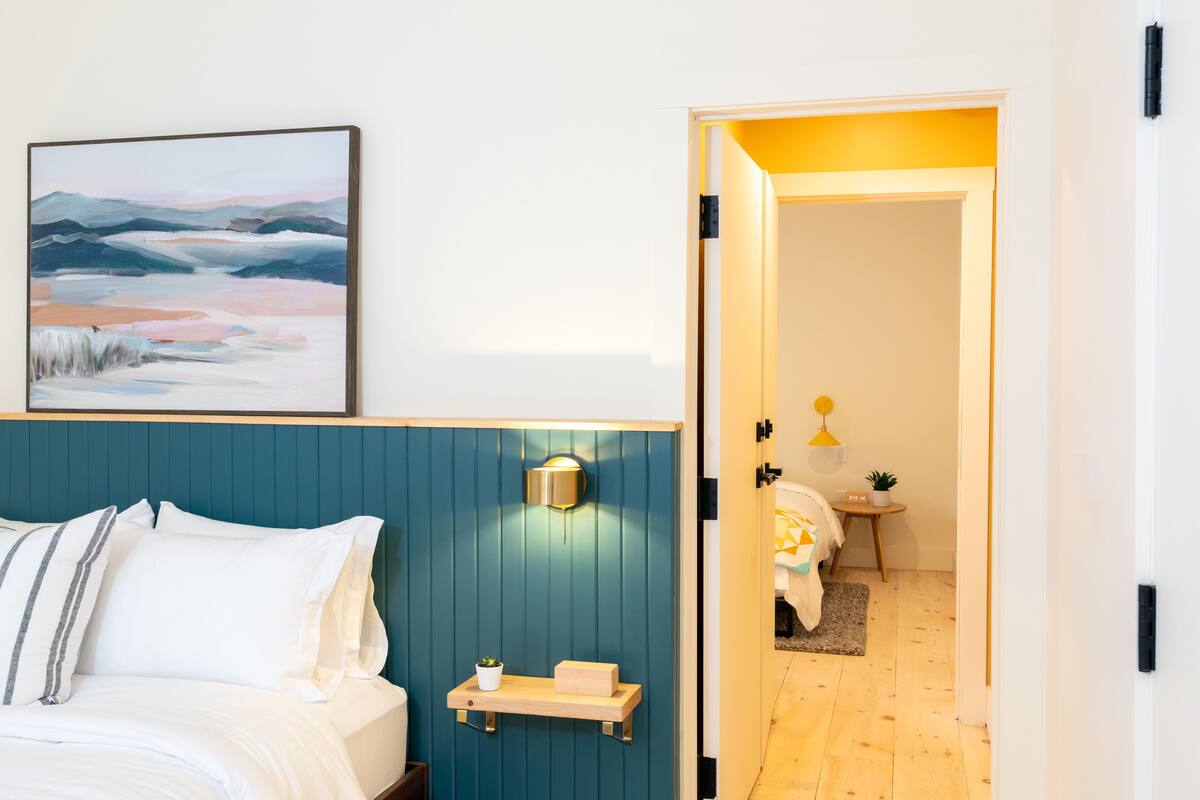 A modern hotel room with a bed, a wall-mounted shelf with a plant, a framed artwork, and a door leading to another room.