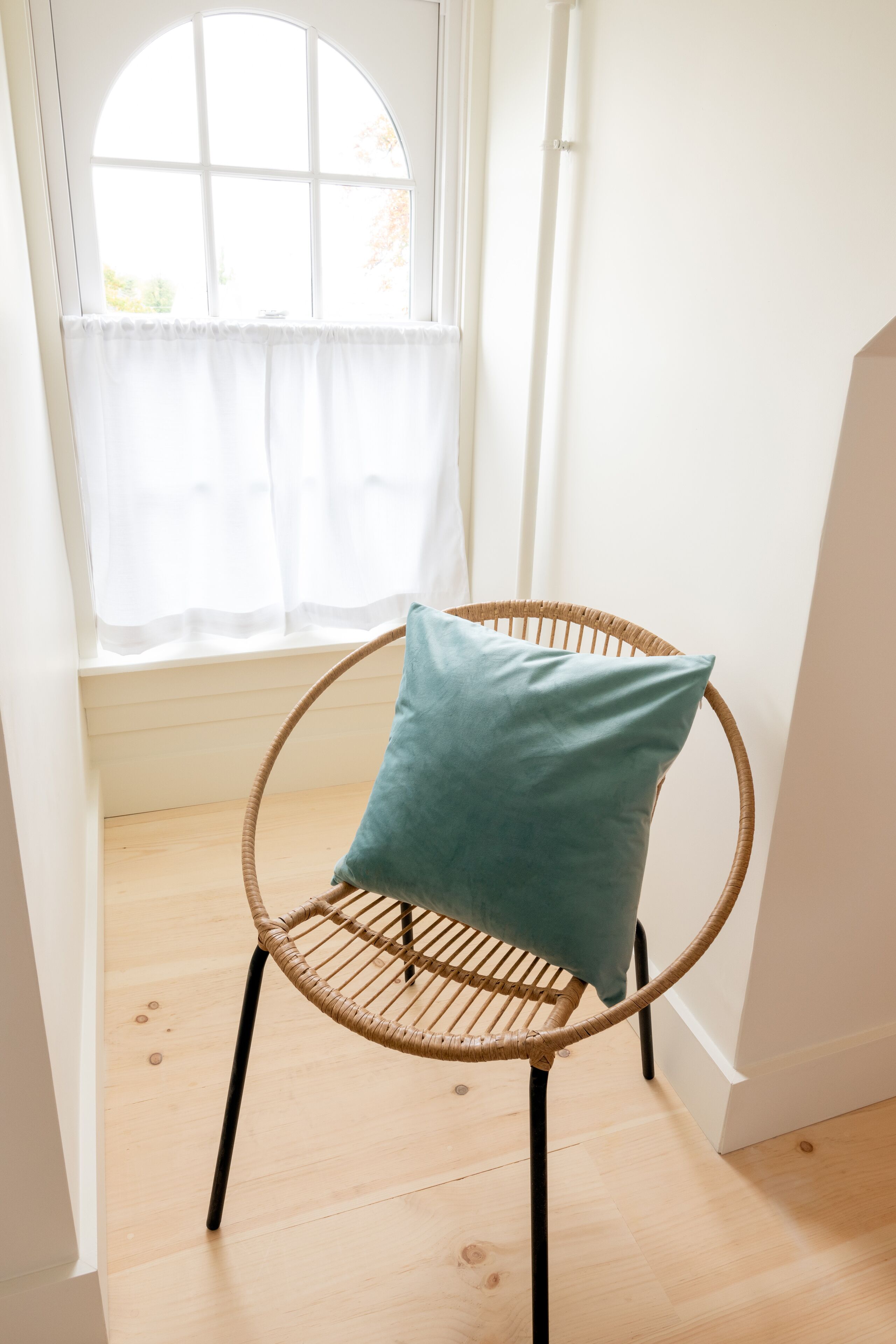 Una silla de mimbre con un cojín azul turquesa en una habitación con un ventanal grande y paredes blancas.