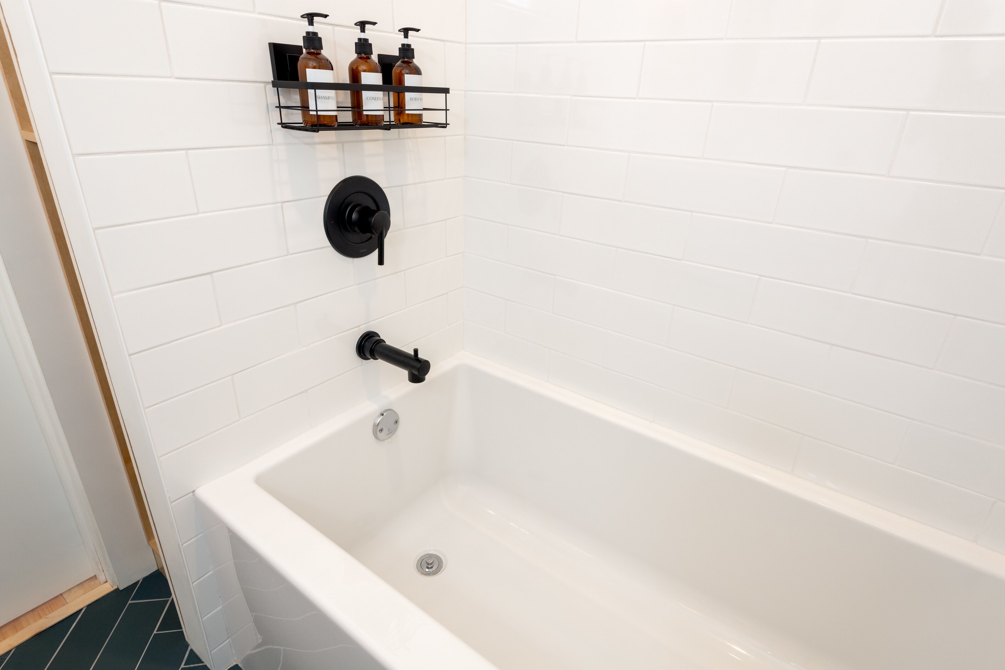 A white bathtub with a black faucet and a chrome drain. There are three bottles on a black shelf above the bathtub.