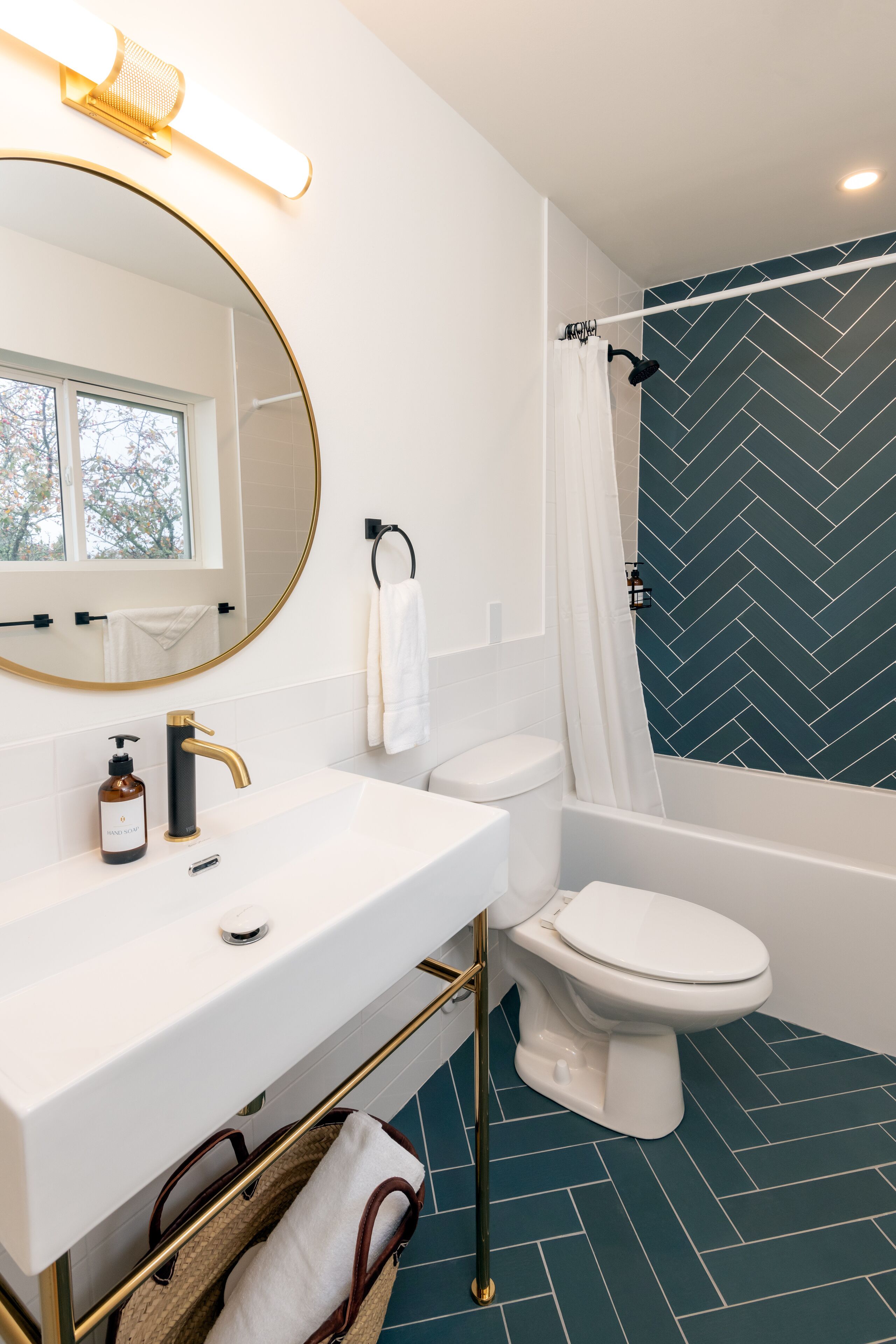 Um banheiro com pia branca, torneira dourada e espelho com moldura dourada. Há um vaso sanitário branco e uma banheira com cortina de chuveiro branca. O chão tem um padrão de mosaico azul em forma de losango.