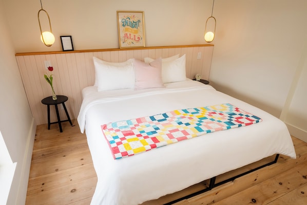 A neatly made bed with a colorful quilt, a wooden headboard, a small bedside table with a vase of flowers, and a framed quote on the wall. - The August House (Windsor)