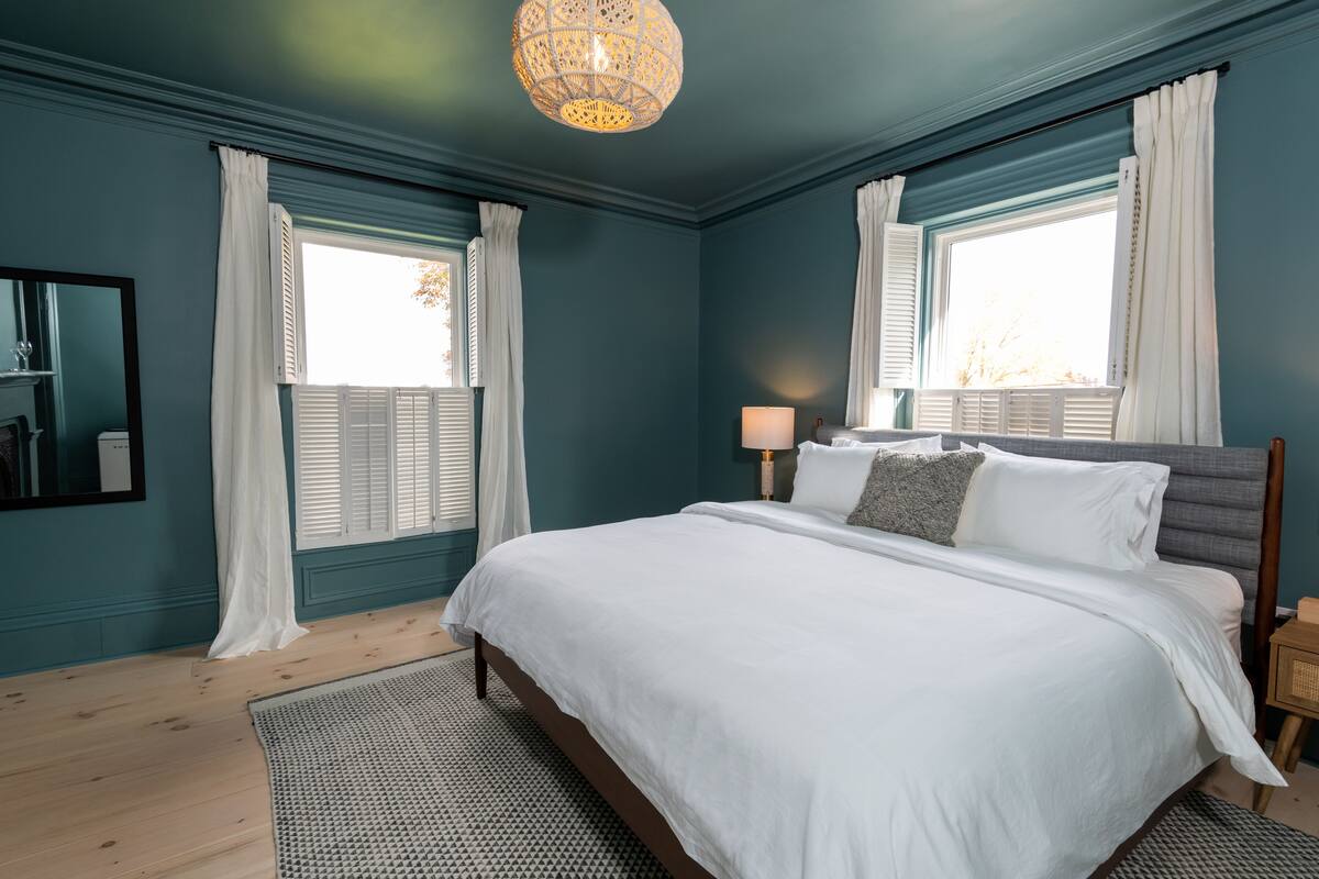 A bedroom with a large bed, two bedside tables, a nightstand, a lamp, and a window with white curtains.