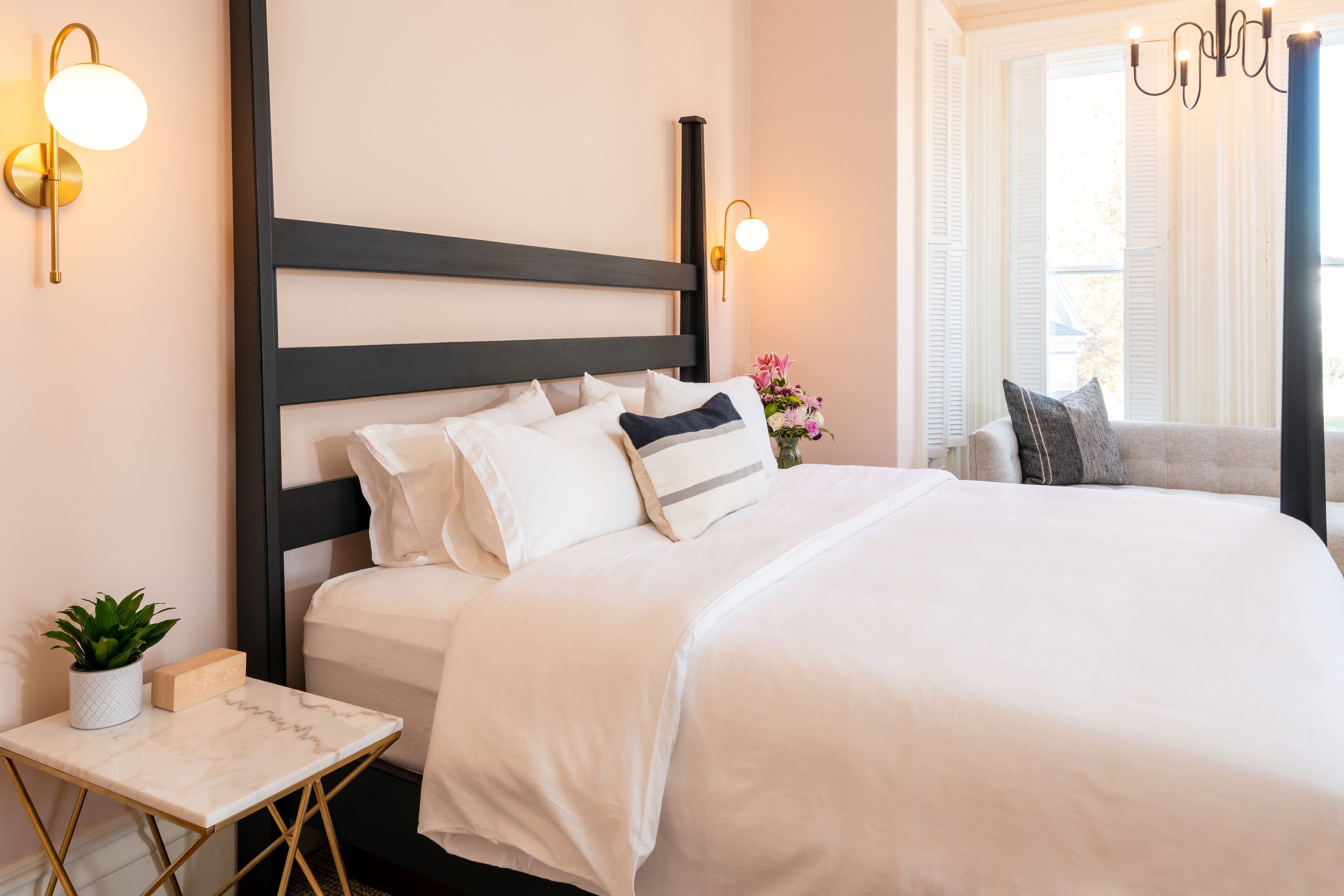 Una cama bien tendida con ropa blanca y varios almohadones, una mesita de noche con una planta y una lámpara, y una ventana con contraventanas blancas.