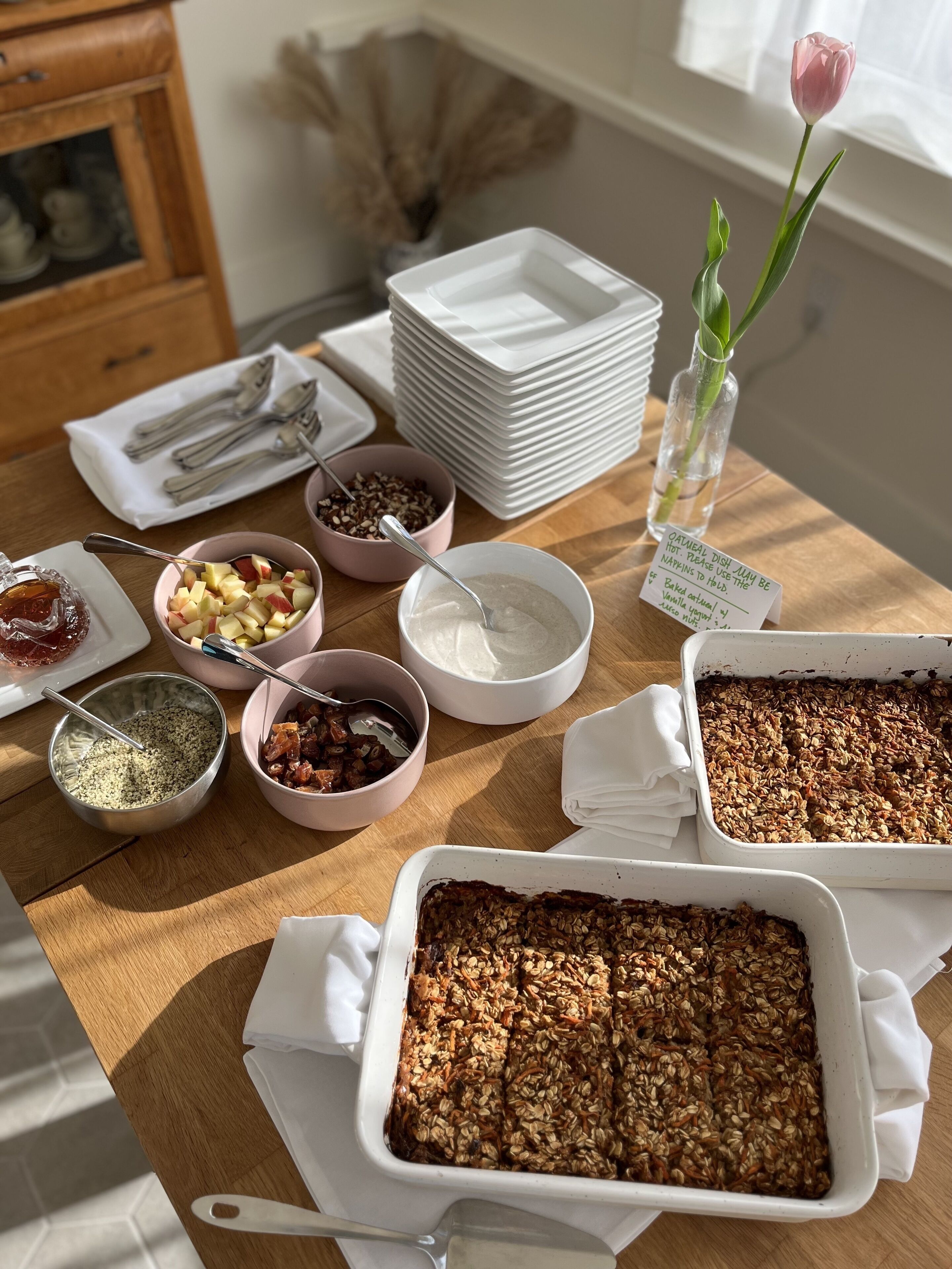 A dining table with various dishes, including a baked dish with oats, bowls of nuts and fruits, and a stack of white plates.
